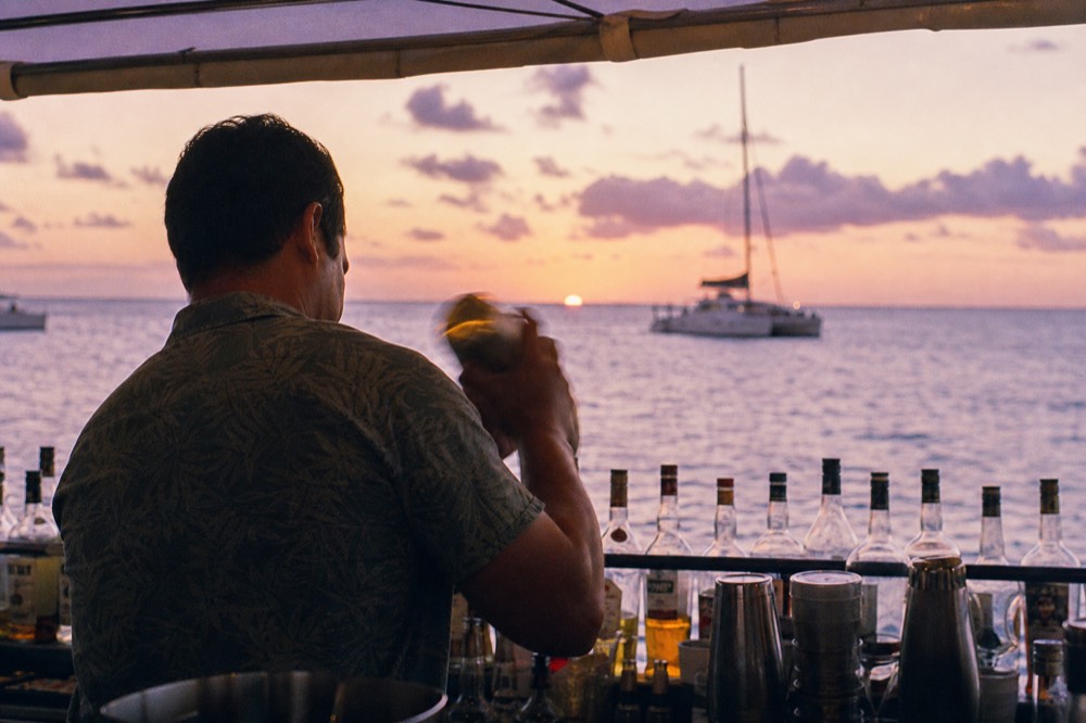 A crew member shaking a cocktail behind a small open-air boat bar, bottles racked against a rail, ocean and low sun blurred beyond