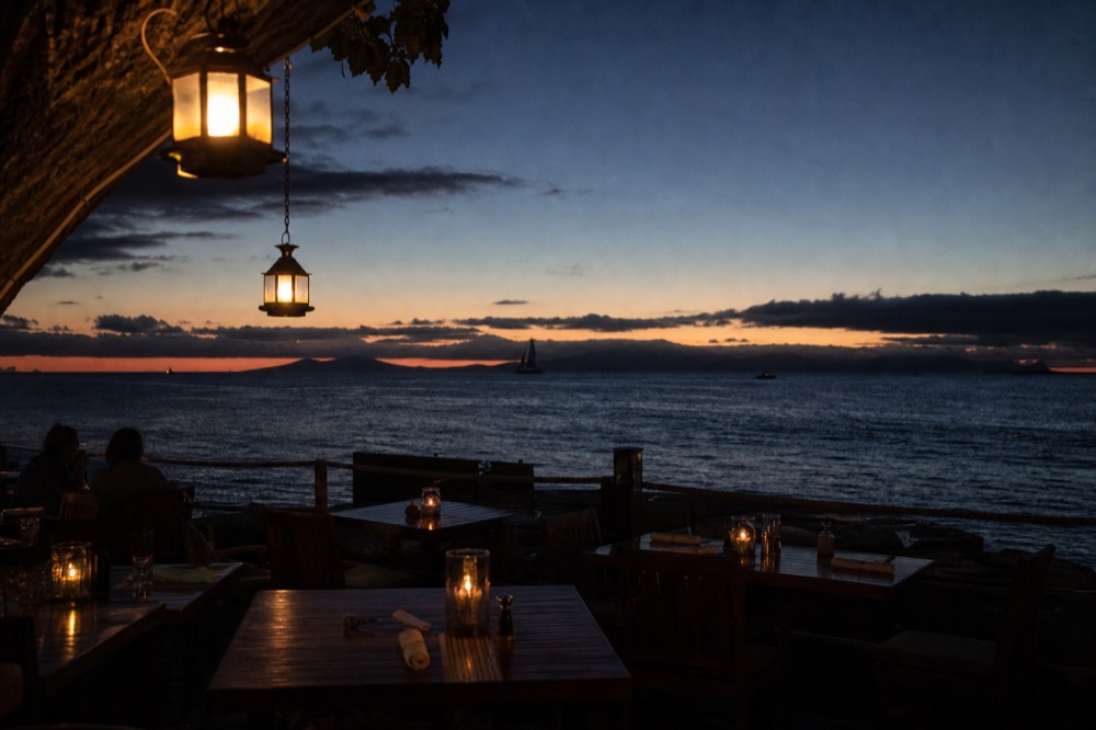 An open-air restaurant terrace at blue hour, lanterns lit over wooden tables, last colour draining from the sky above a dark ocean horizon