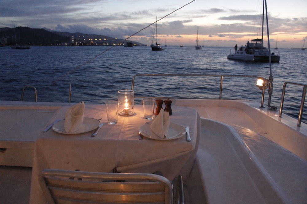 An empty white-linen table set for two on the aft deck of a catamaran, a candle lantern lit, water darkening beyond the rail