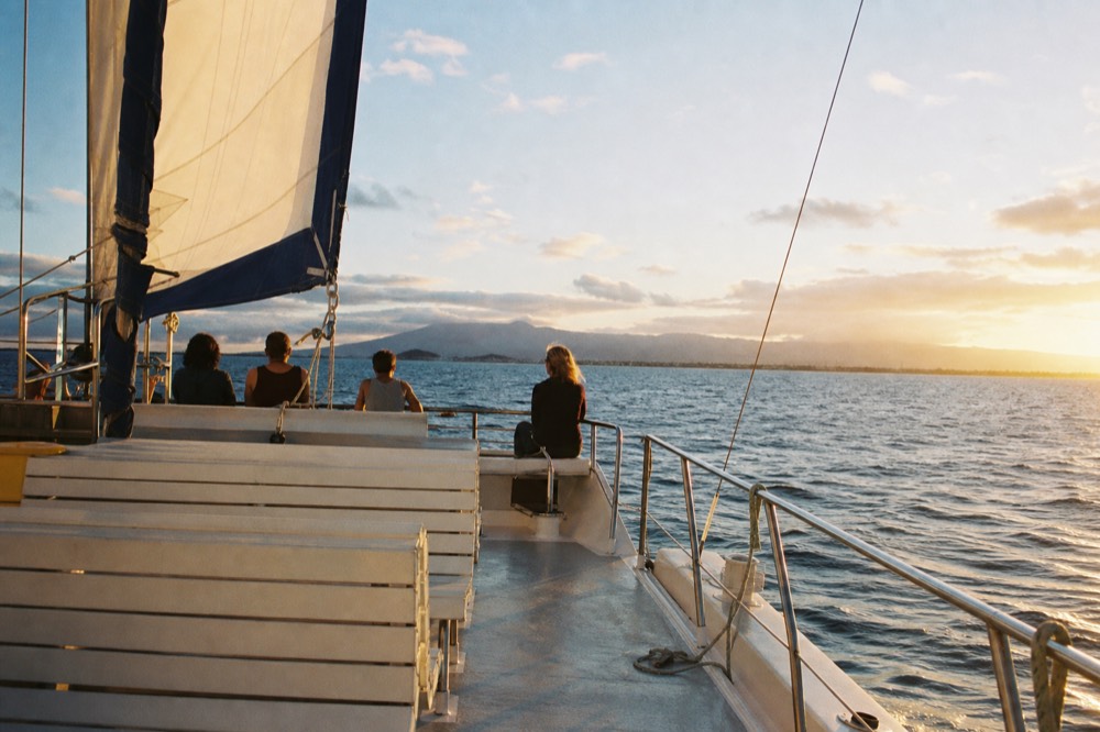 A wide catamaran deck with only a few guests seen from behind at the rail, plenty of open bench space, warm golden light