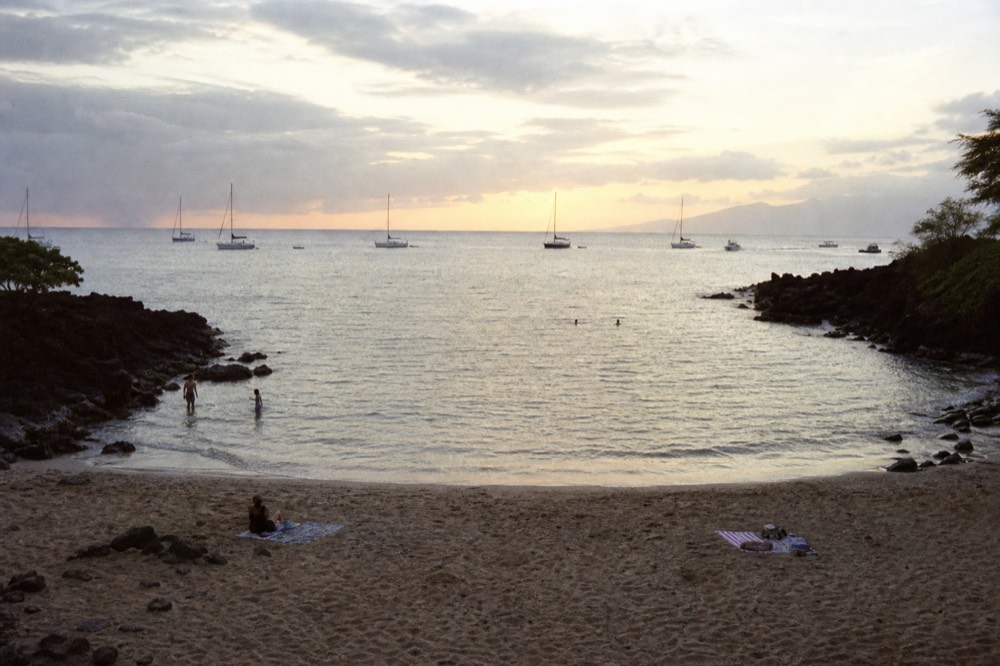 A small crescent cove at dusk with lava points on both sides framing the last light, calm water inside the bay, a few beach towels on the sand