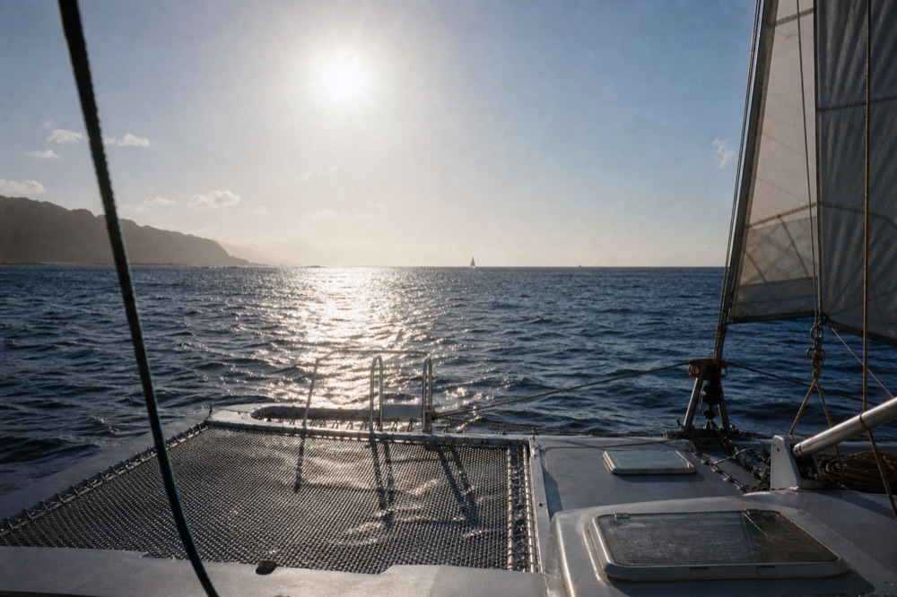 View forward from a catamaran deck about an hour before sunset, sun still high and white over open channel water, sails half raised