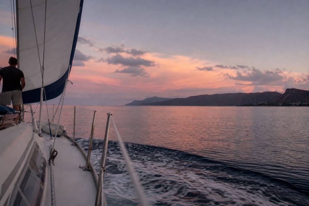 A catamaran under sail in a channel at golden hour, long island ridge on the horizon ahead, wake trailing across smooth water