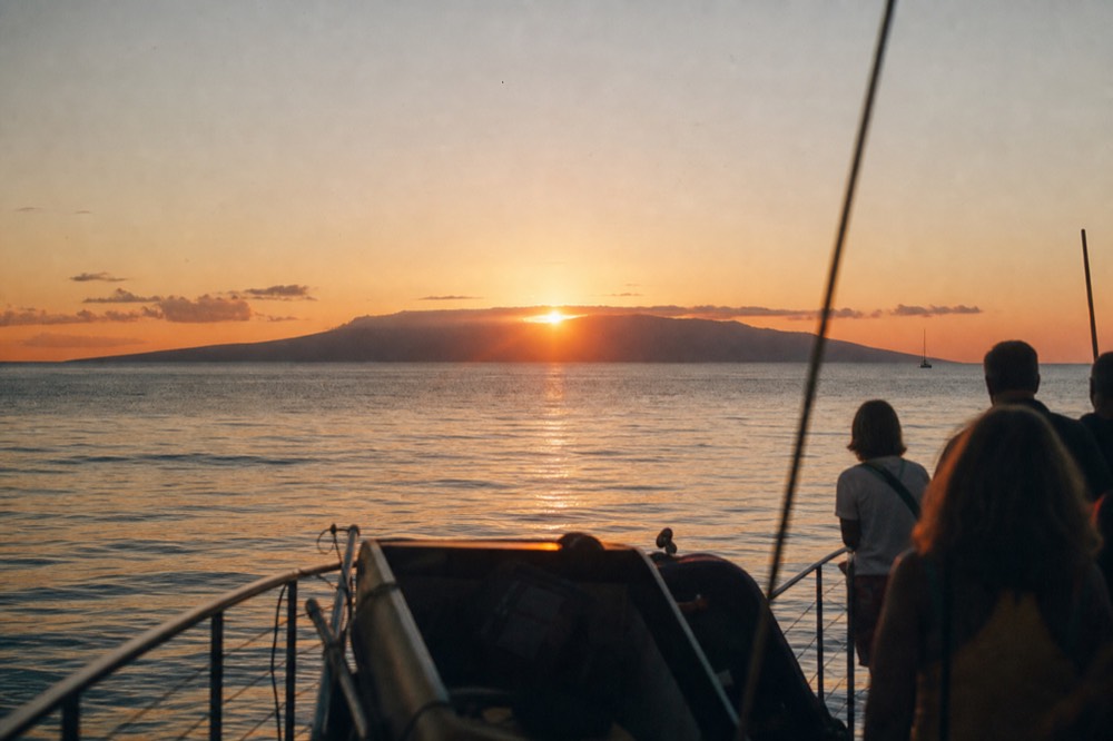 Sunset seen from a boat deck with a long low island profile dead ahead, sun edge just touching its ridge, glassy water reflecting orange