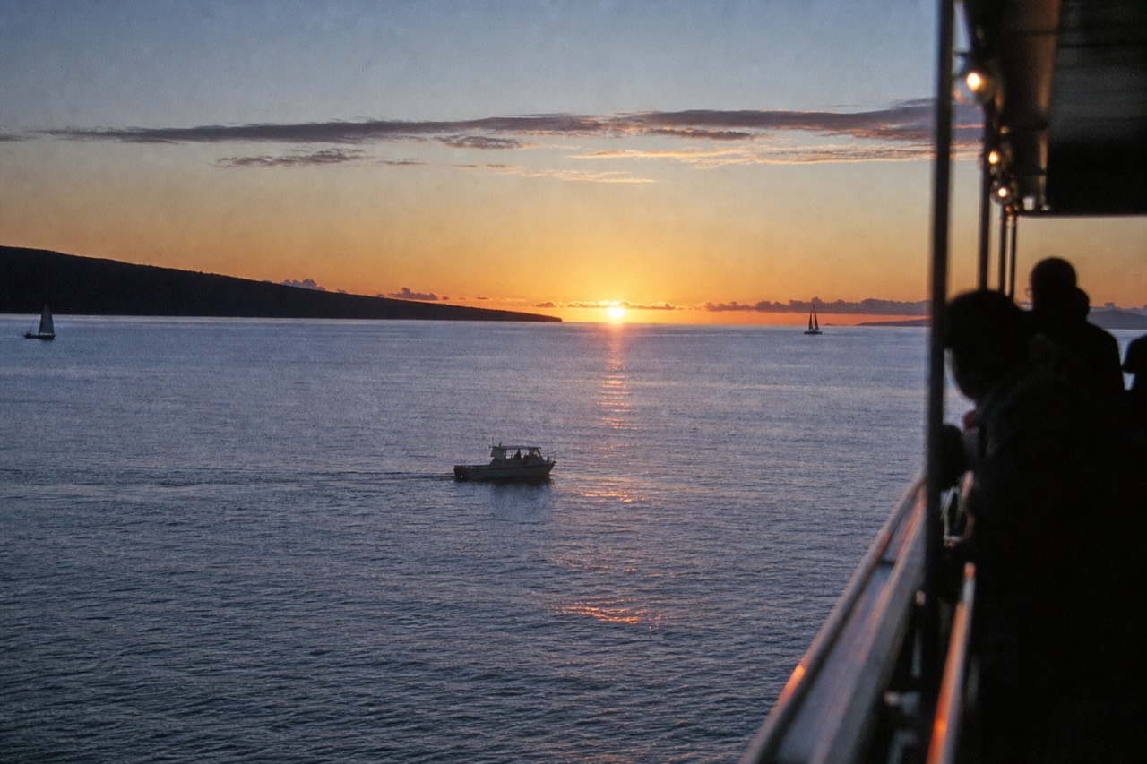 The sun low over a calm channel between two islands, one long low island profile dark against orange sky, the water almost without swell
