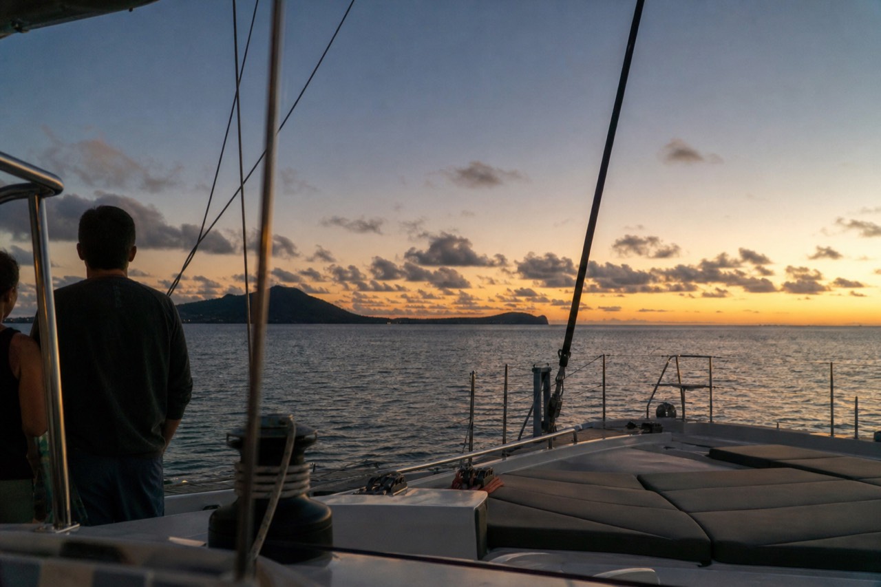 A wide catamaran deck at golden hour with flat green channel water behind it and a low island shape on the horizon