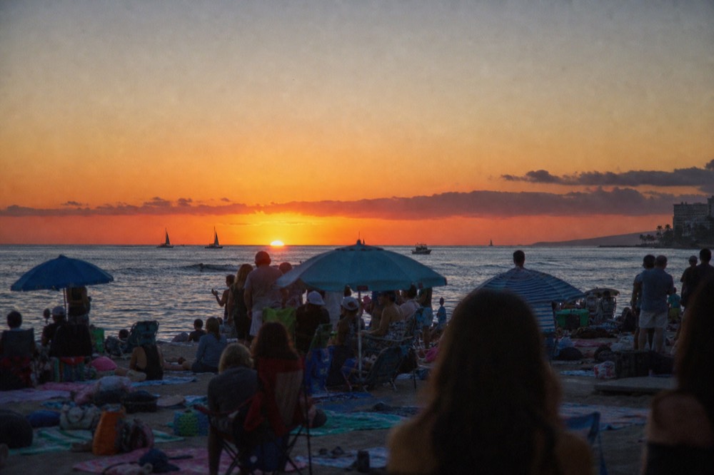 A crowded west-facing beach at dusk with dozens of towels, umbrellas and standing figures blocking the low sun