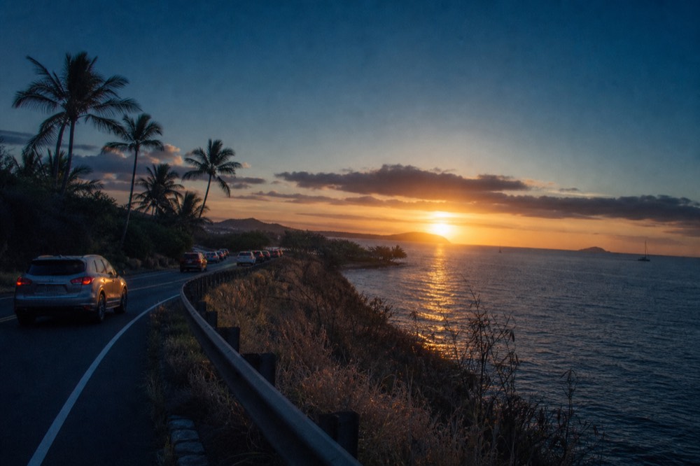 Two-lane coastal highway curving along a dry cliff above the ocean with low sun ahead and a line of cars in the distance