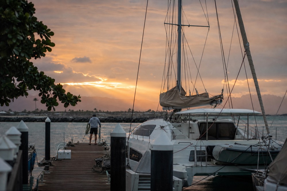 Wooden harbor dock with numbered slip posts leading toward a moored catamaran and a crew member coiling a line at dusk
