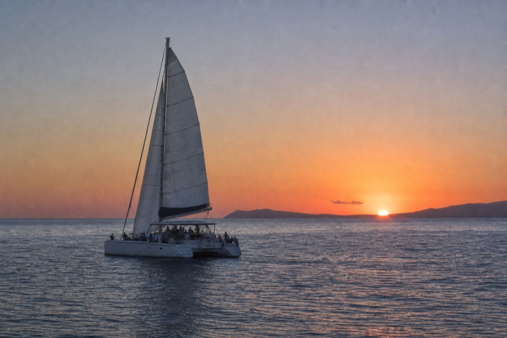 Sailing catamaran under full white sail on flat evening water with a long low island ridge on the horizon