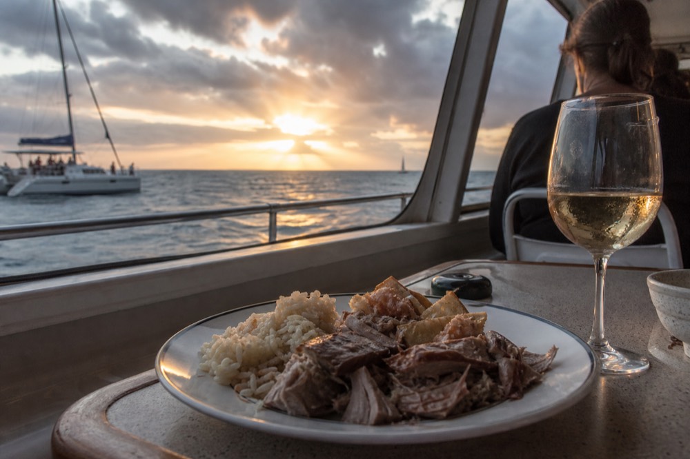 Plate of carved meat and rice on a boat table with a wine glass catching orange light from the window beside it