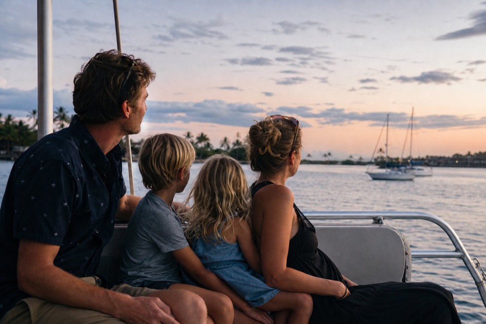 Family of four seated together on a boat bench at sunset with soft light on their faces turned toward the water
