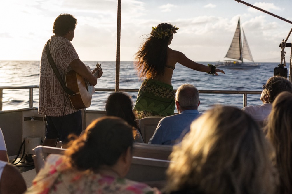 Guitarist and a hula dancer performing on an open boat deck at dusk with seated guests watching
