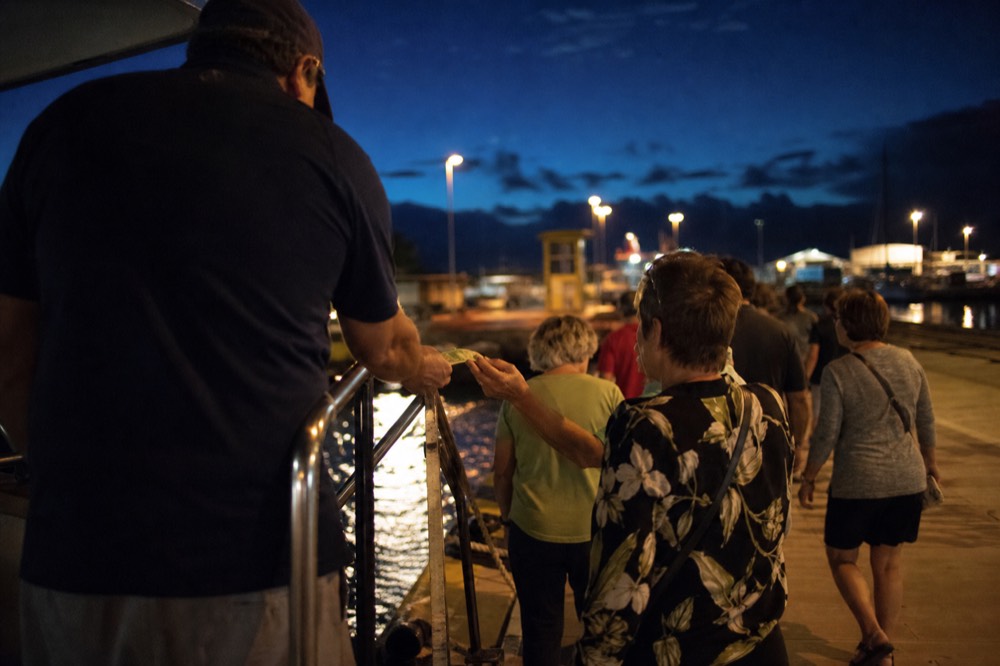 Guests handing cash to a crew member at a gangway rail as they step off a boat under harbor floodlights after dark
