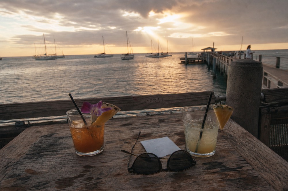 Overhead shot of a small paper receipt, a pair of sunglasses and two half-finished tropical drinks on a weathered wooden dock table