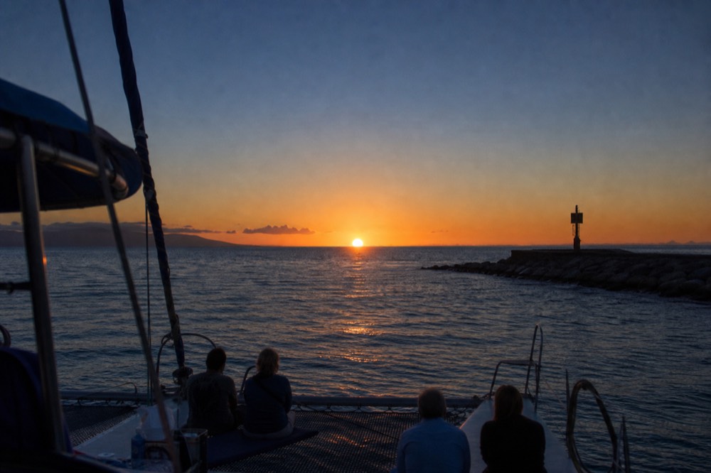 Small open-bar catamaran with only a handful of guests aboard heading out past a harbor wall in late afternoon light