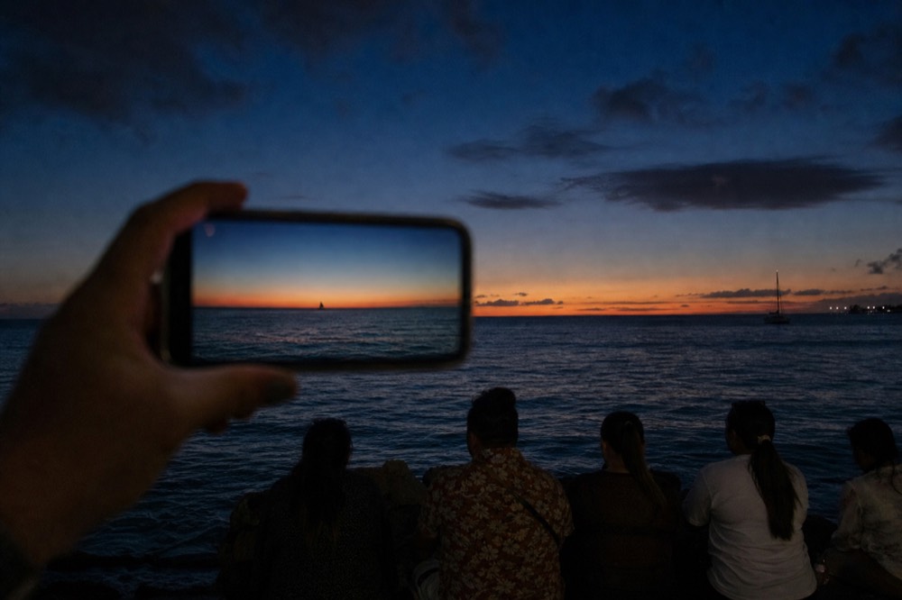 Phone screen held up against a wide ocean horizon at sunset showing how small a distant dark shape appears in frame