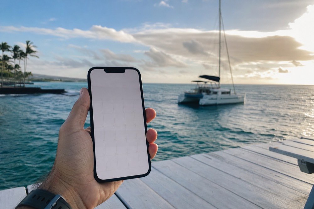 Hand holding a phone showing a blank booking calendar while a catamaran waits offshore in soft evening light