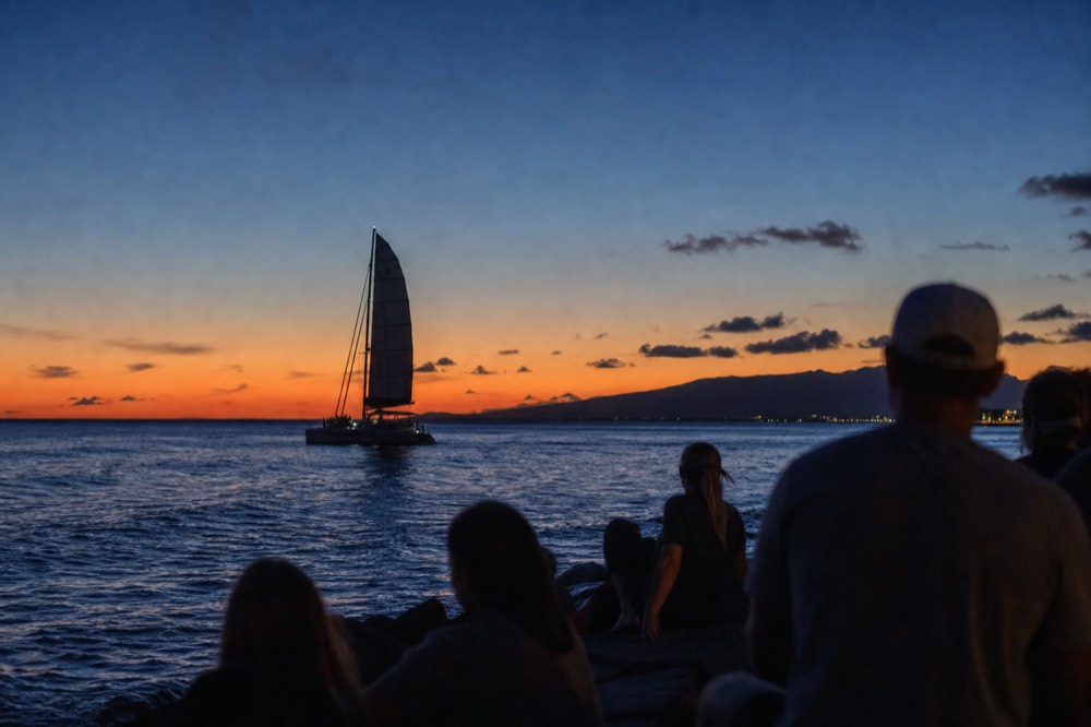 Catamaran silhouette crossing the last band of orange light on the water with a mountainous island shape behind it at blue hour