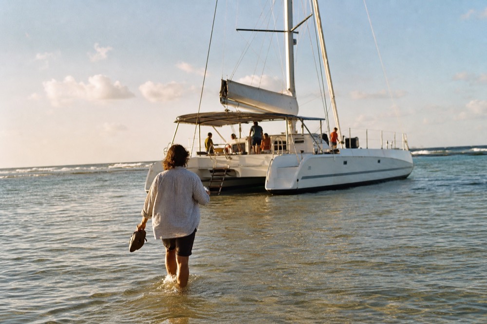 Person wading shin-deep through calm shallows toward a catamaran with sandals held up in one hand
