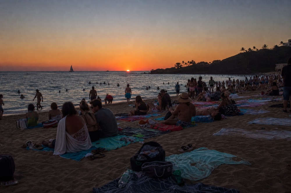 Crowded golden sand beach half an hour before sunset with towels overlapping and a dark lava headland at the far end