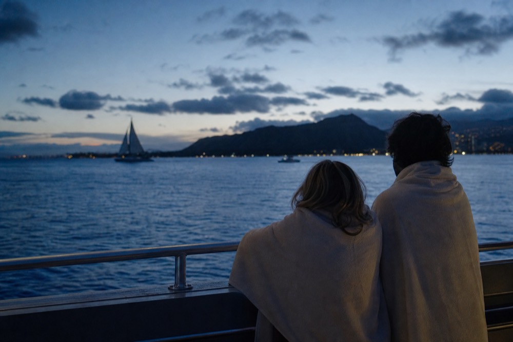 Two people at a boat rail after sunset in blue-hour light with a warm layer over their shoulders and shoreline lights behind them